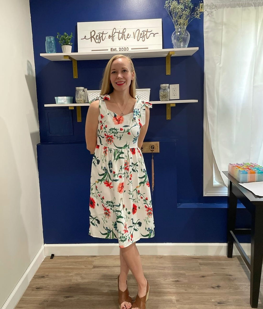 Kellie from Rest of the Nest Keepsake Jewelry Store, wearing a floral dress with a blue wall behind her with filled with shelves, pictures and trinkets